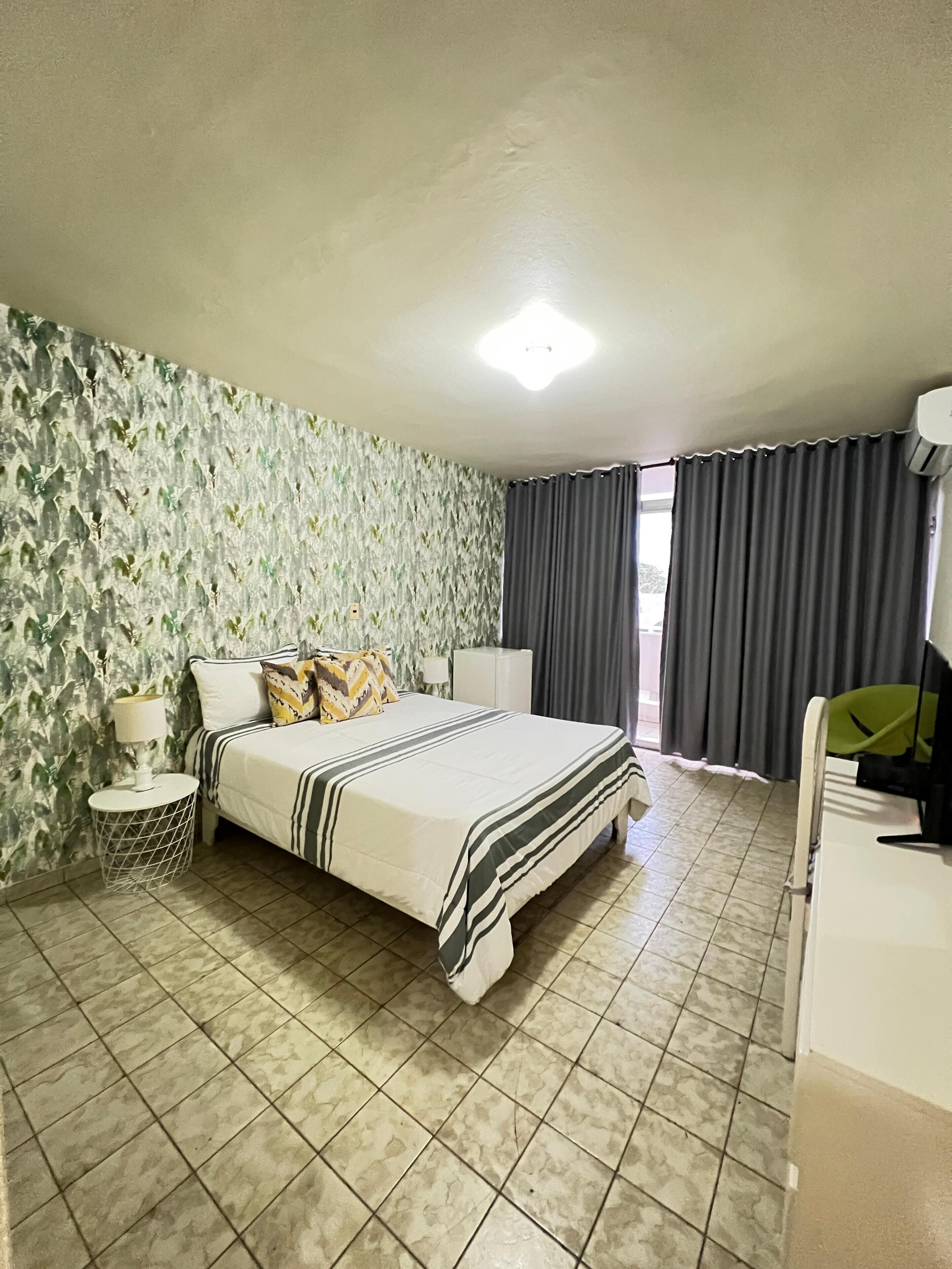 Une chambre d’hôtel avec un lit, une table de chevet avec une lampe, une commode et une fenêtre avec des rideaux.