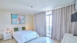 A hotel room with a bed, a nightstand, a lamp, a painting on the wall, and a view through the curtains. - Modern Santo Domingo Condo in Heart of Gazcue (Santo Domingo)