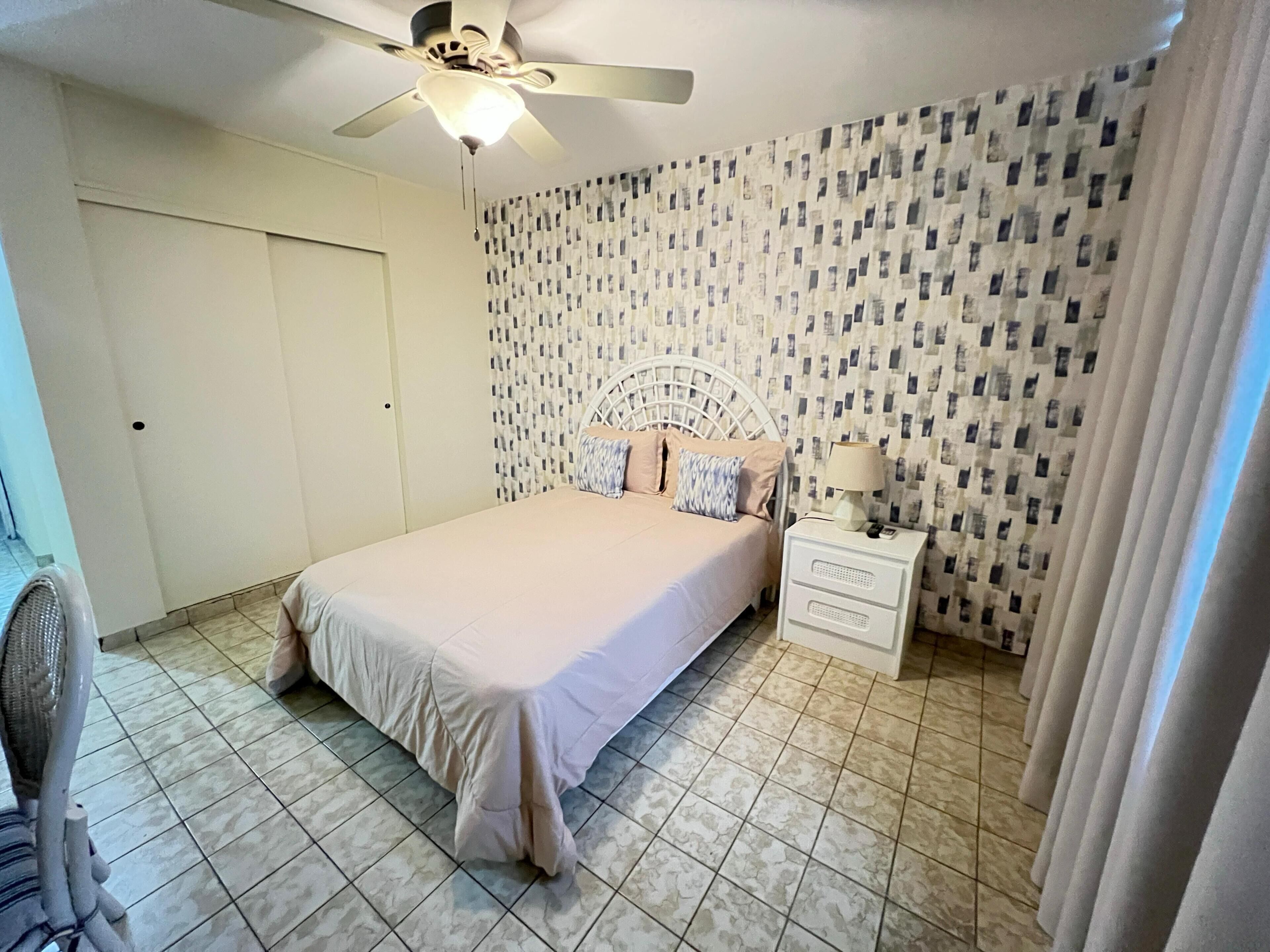 A bedroom with a bed, a nightstand, a ceiling fan, and patterned wallpaper.