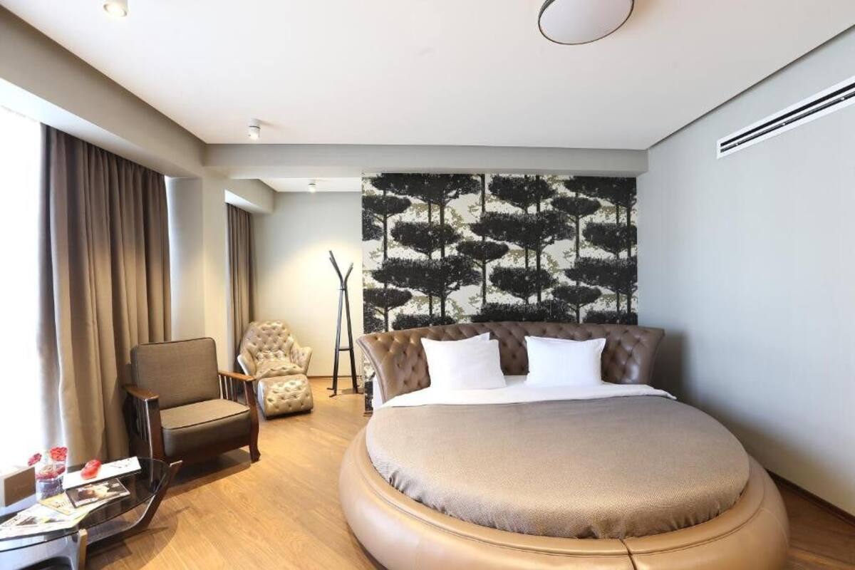 A modern hotel room with a round bed, a seating area with a chair, a small table, and a large wall mural.