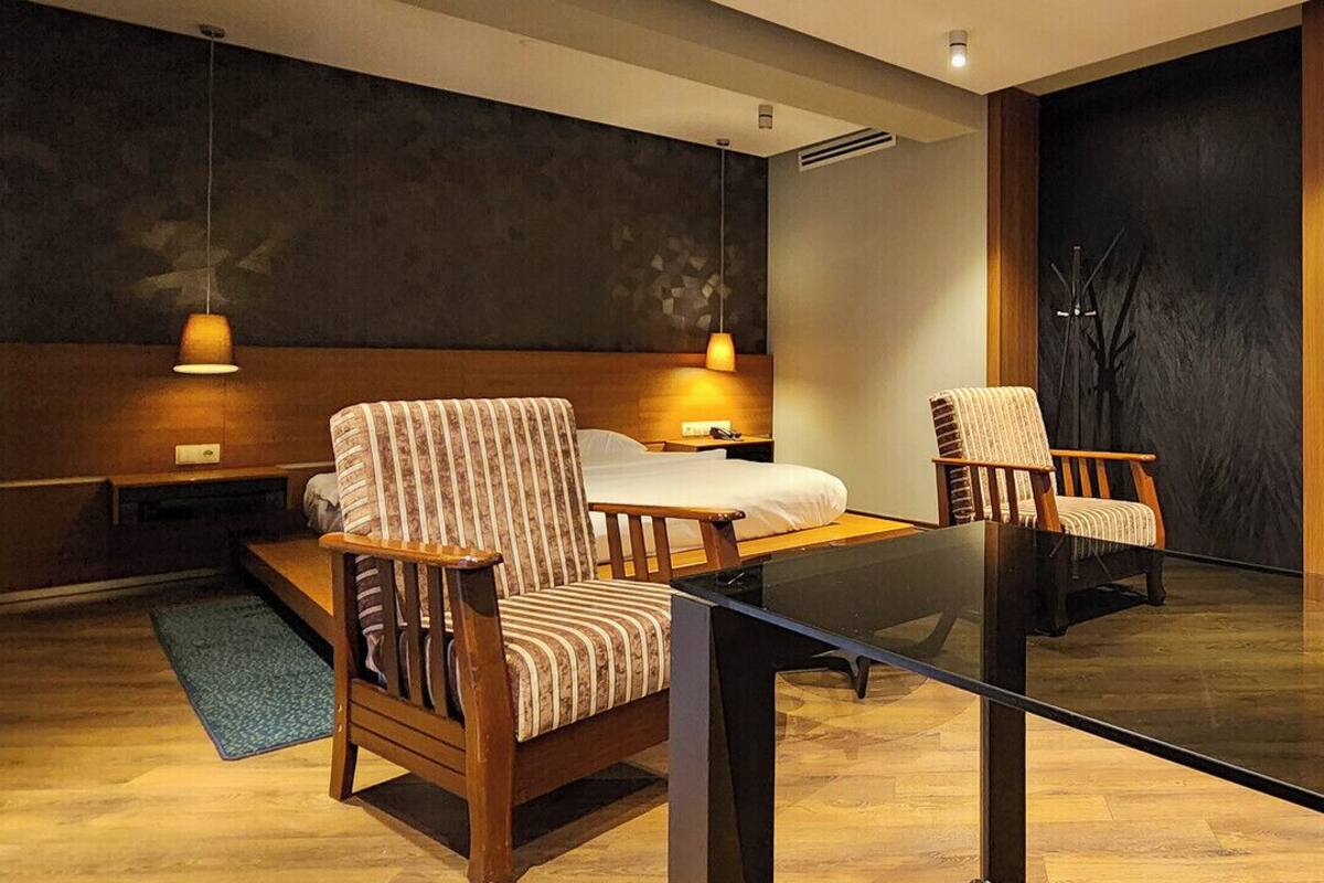 A modern hotel room with a large bed, two upholstered chairs, a glass coffee table, and a dark accent wall.