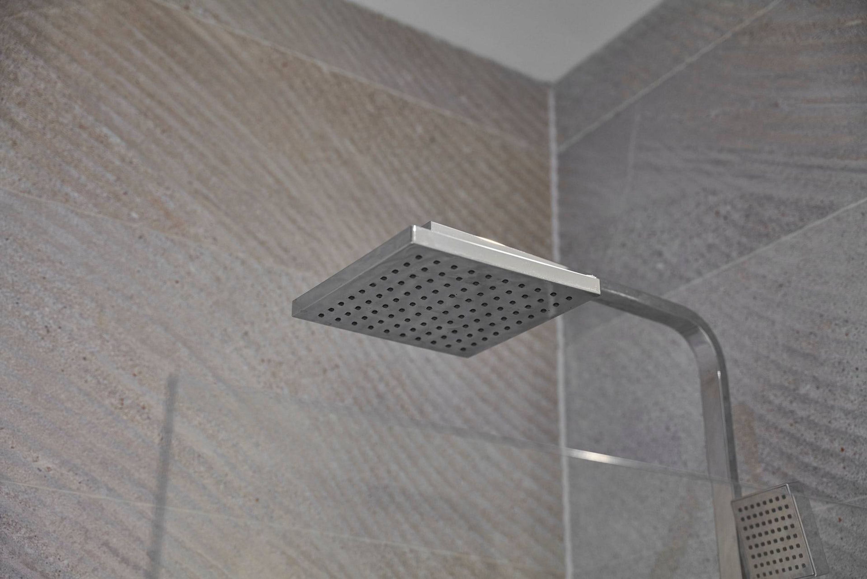 A showerhead with a square, perforated design mounted on a curved arm, set against a textured wall.
