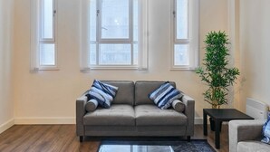 Ein Wohnzimmer mit grauem Sofa, zwei Fenstern, einer Pflanze und einem Glastisch.