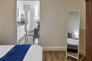 A hotel room with a bed, a blue towel, a wooden floor, a mirror, and a view of a kitchen and dining area. - Host Stay Three Graces Getaway (Liverpool)
