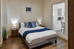 A bedroom with a bed, bedside tables, a nightstand, a potted plant, and a view of a kitchen through a glass door. - Host Stay Three Graces Getaway (Liverpool)