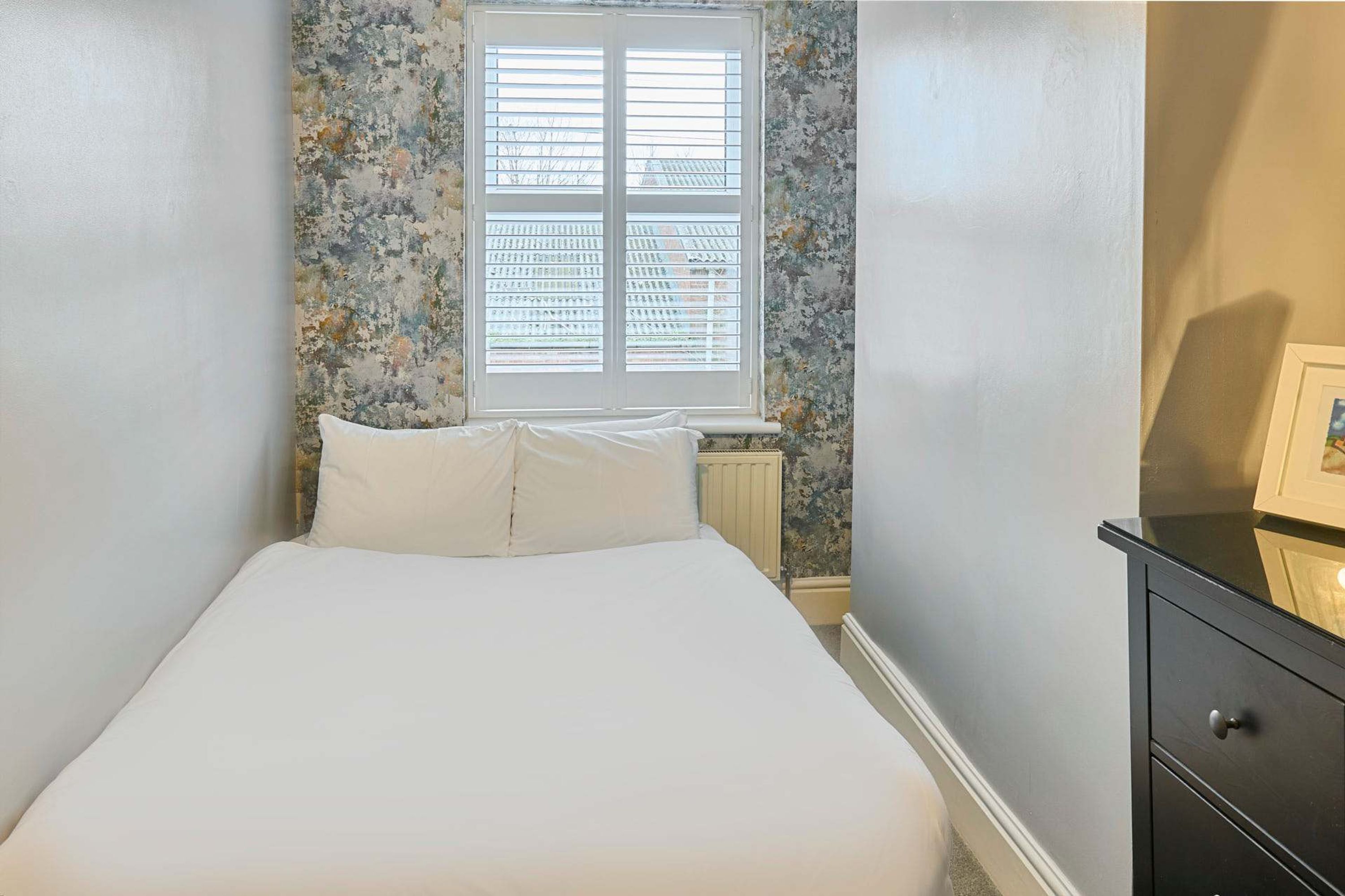 Ein Schlafzimmer mit Bett, Fenster mit Jalousien, Heizkörper und Kommode.