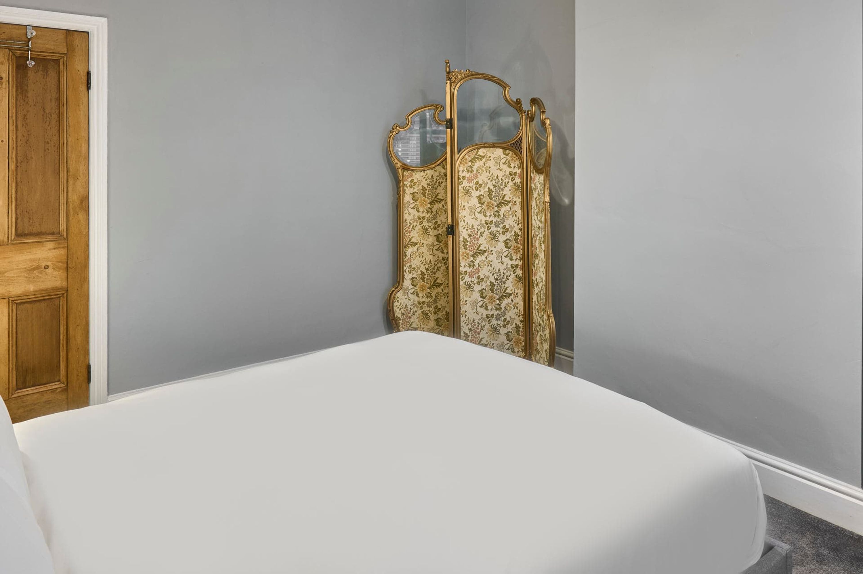 A bedroom with a wooden door, a white bed, and a decorative mirror with a floral pattern.
