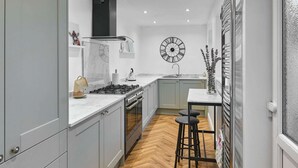 A compact kitchen with grey cabinets, a white countertop, a central stove, and a sink. A large wall clock and a decorative plant are also visible. - Host Stay Railway Cottages (Liverpool)