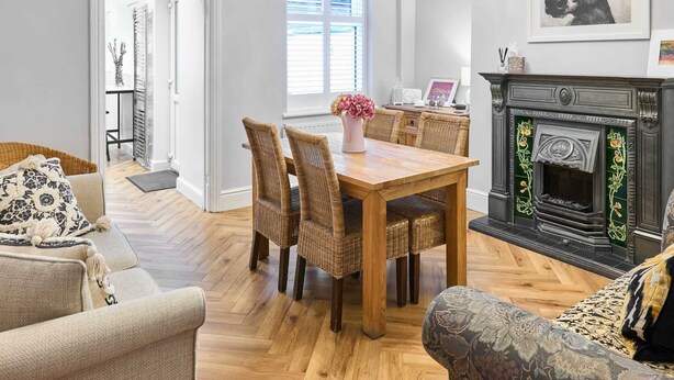 A dining area with a wooden table and wicker chairs, a fireplace with decorative elements, and a large window with blinds. - Host Stay Railway Cottages (Liverpool)