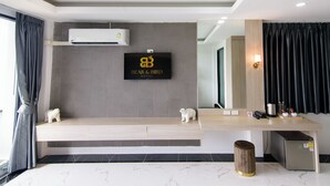 A modern hotel room with a mounted TV displaying the hotel name, a built-in air conditioner, a wooden vanity with a mirror, and a small trash bin. - Bear & Bird Hotel   (Surat Thani)