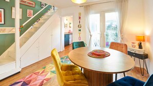 Ruokailutilassa on puinen pöytä, keltaiset ja siniset tuolit, värikäs, geometrisesti kuvioitu matto sekä valkoisella kaiteella varustetut portaat.
