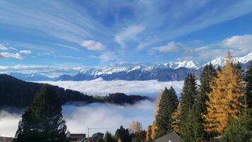 Eine gebirgige Landschaft mit schneebedeckten Gipfeln, einem bewaldeten Tal und einem kleinen Gebäude mit schrägem Dach.
