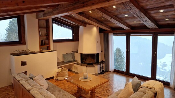 Ein gemütliches Wohnzimmer mit Kamin, Holzbalken, einer Couch und einem großen Fenster mit Bergblick.