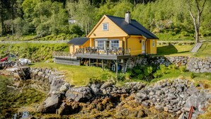 En gul hytte med terrasse, omgivet af en klippefyldt kystlinje og vegetation.