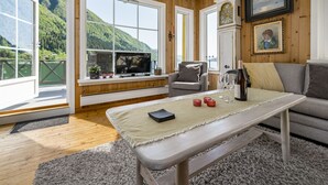 Et hyggeligt værelse med træpaneler, store vinduer der viser udsigt over grønne bakker, et fjernsyn, en sofa og et bord med en flaske og glas.