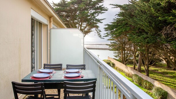 Un balcón con una mesa lista para dos personas, con vistas al agua y a una zona boscosa.