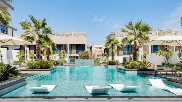 Ett modernt poolområde med en klarblå pool, vita solstolar, palmer och en dekorativ vattenfallsanläggning.