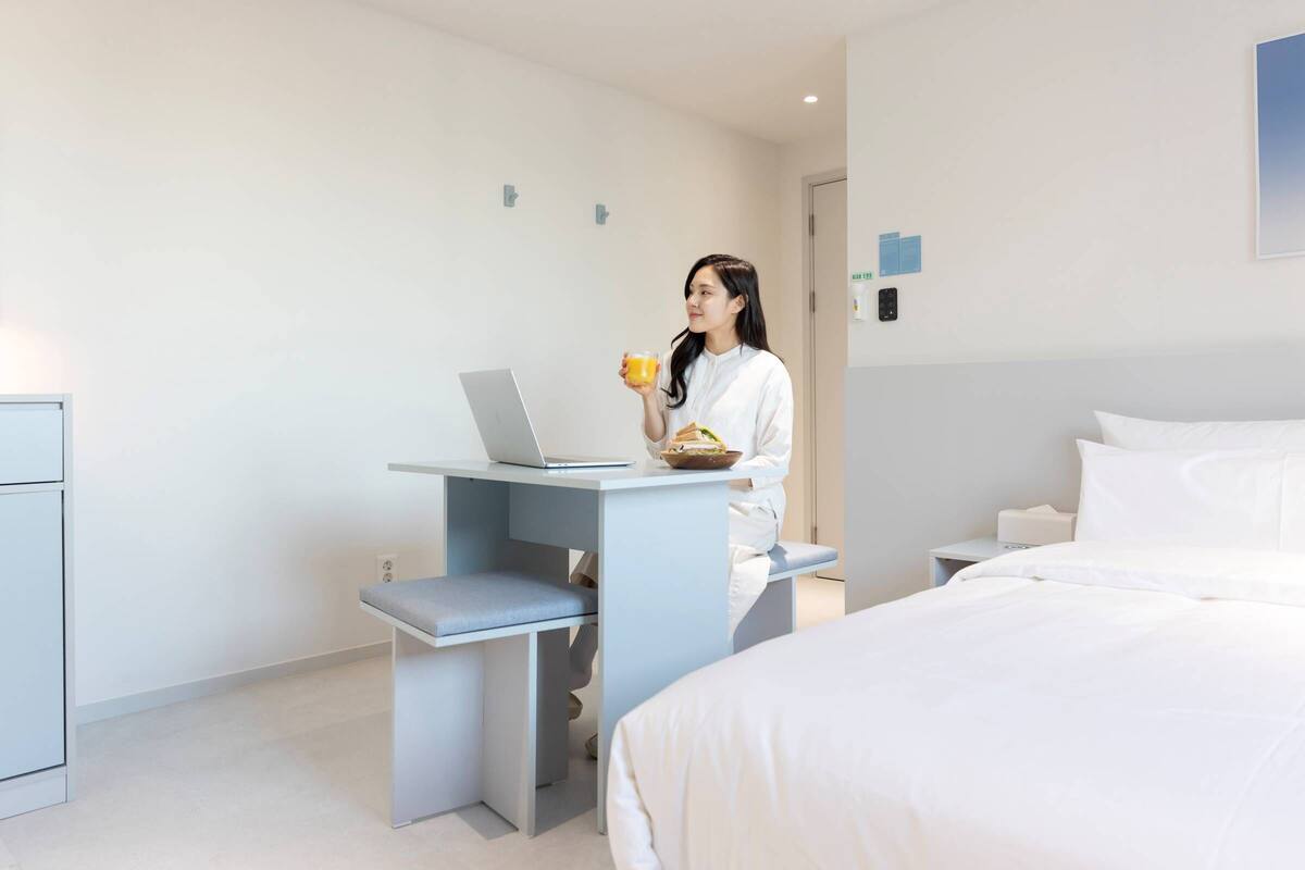 Une chambre d’hôtel moderne avec un lit, un bureau doté d’un ordinateur portable, et une personne tenant une tasse.