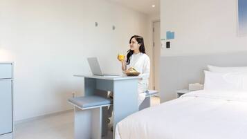 A modern hotel room with a bed, a desk with a laptop, and a person holding a cup.