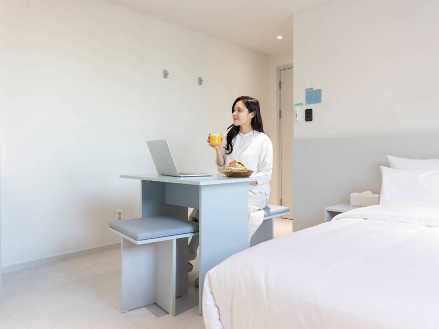 A modern hotel room with a bed, a desk with a laptop, and a person holding a cup.