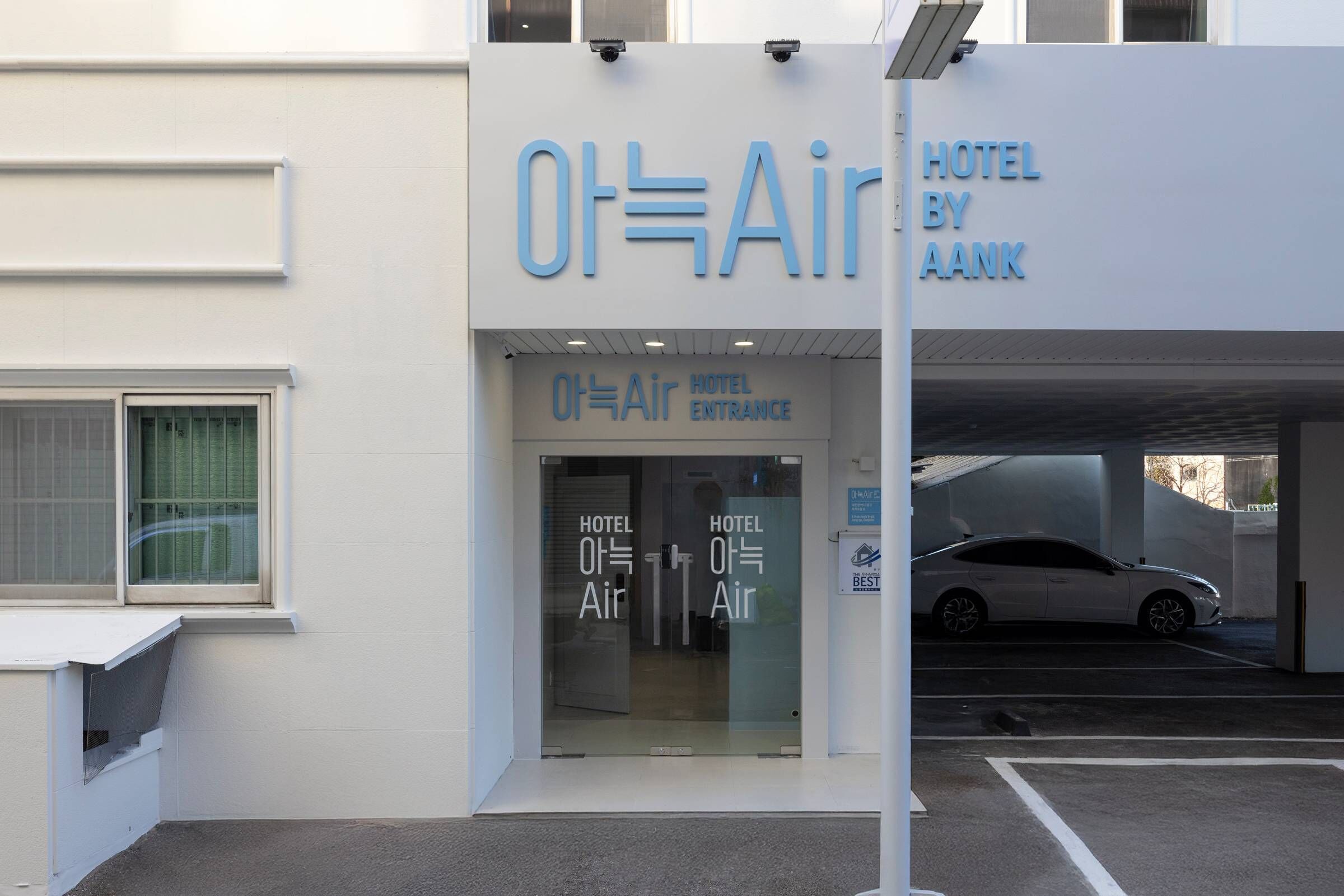 La entrada al hotel Ot=Air by AANK, con un diseño moderno y fachada de vidrio, además de un estacionamiento visible.
