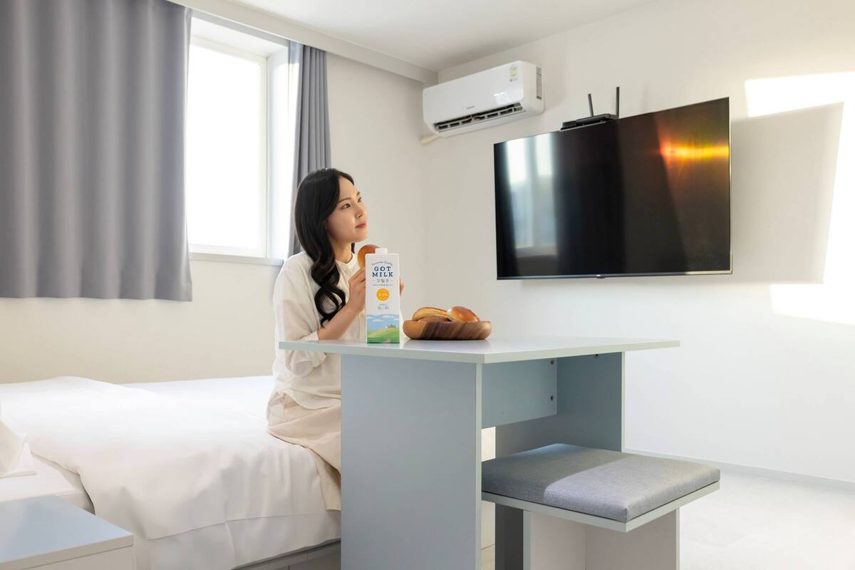 Une chambre d’hôtel avec un lit, une petite table sur laquelle se trouve un bol de pain, une bouteille de lait et un téléviseur fixé au mur.
