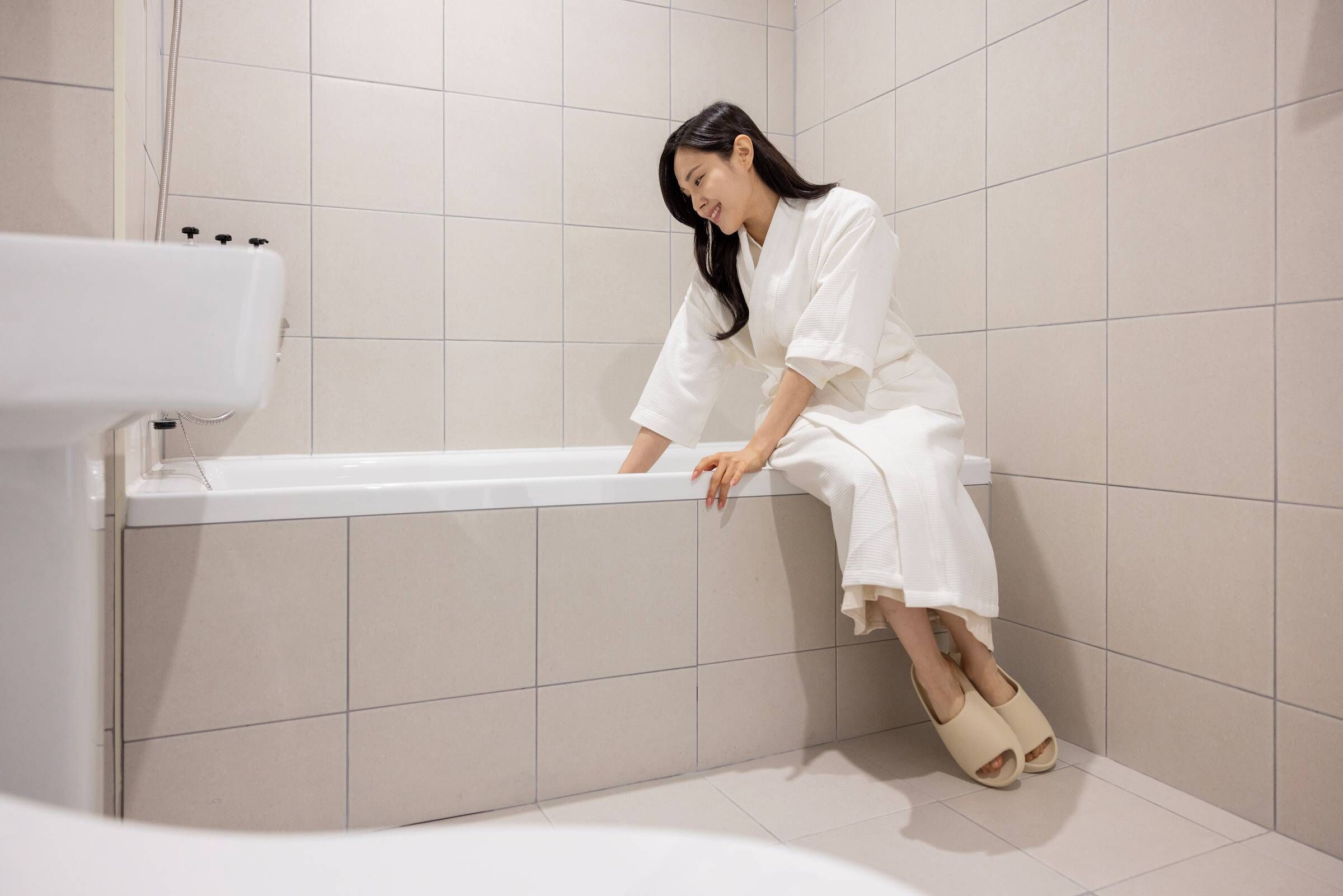 A person sitting on the edge of a bathtub in a bathroom with tiled walls and floor.