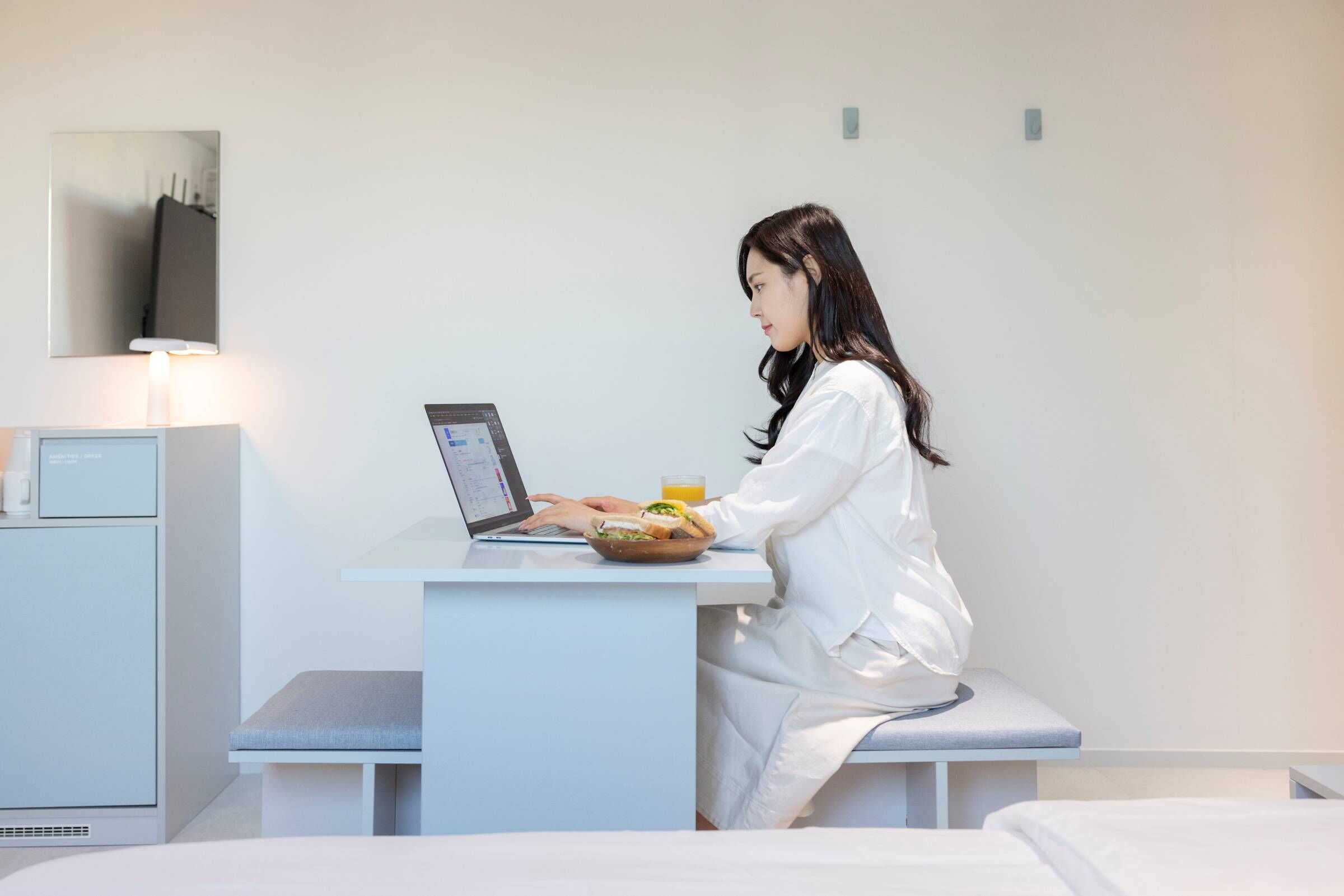 A modern hotel room with a bed, a desk with a laptop, and a small table with a sandwich and a glass of orange juice.