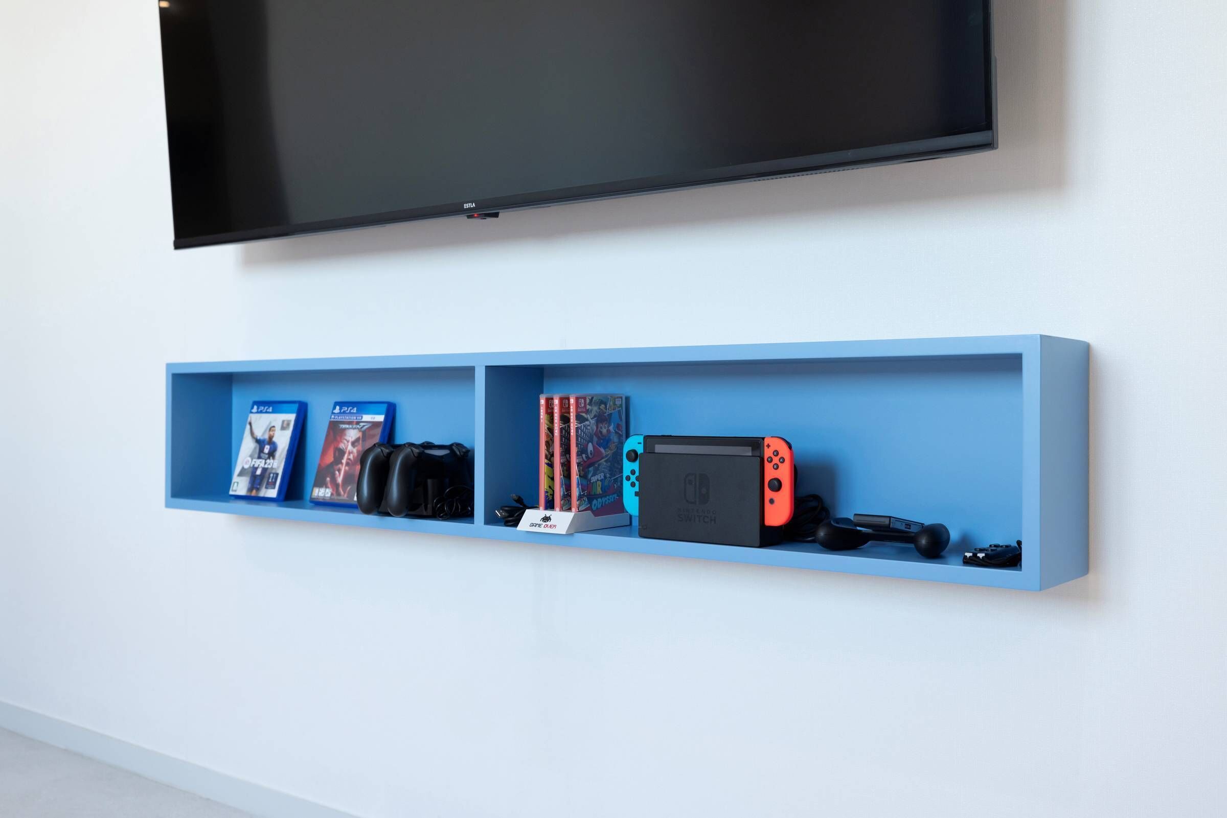 Un estante azul con videojuegos, una Nintendo Switch y un televisor montado en una pared blanca.