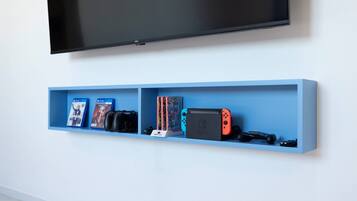 Un estante azul con videojuegos, una Nintendo Switch y un televisor montado en una pared blanca.