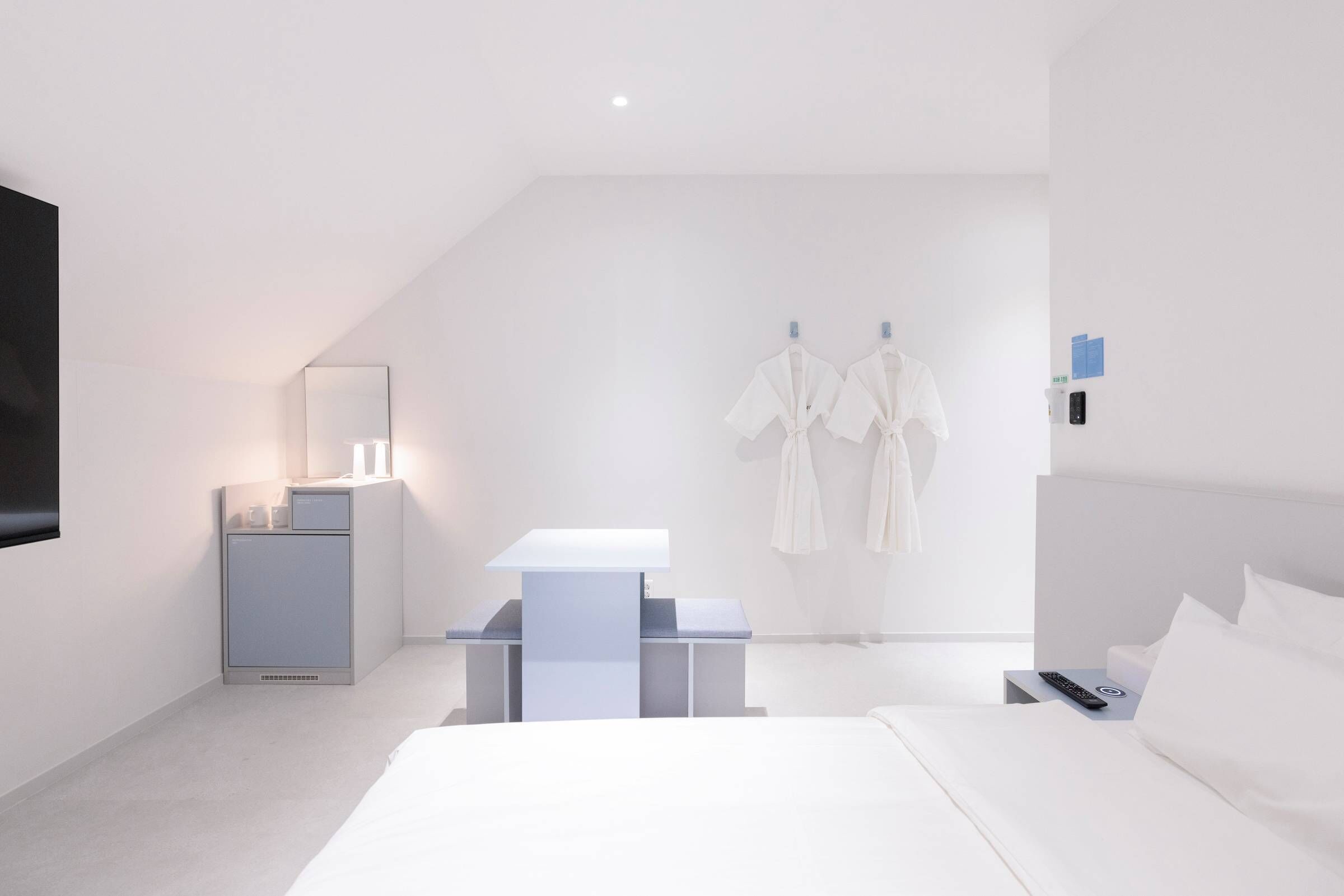 A modern hotel room with a bed, bedside table, and a small desk with a chair. There is a TV mounted on the wall and a window providing natural light.