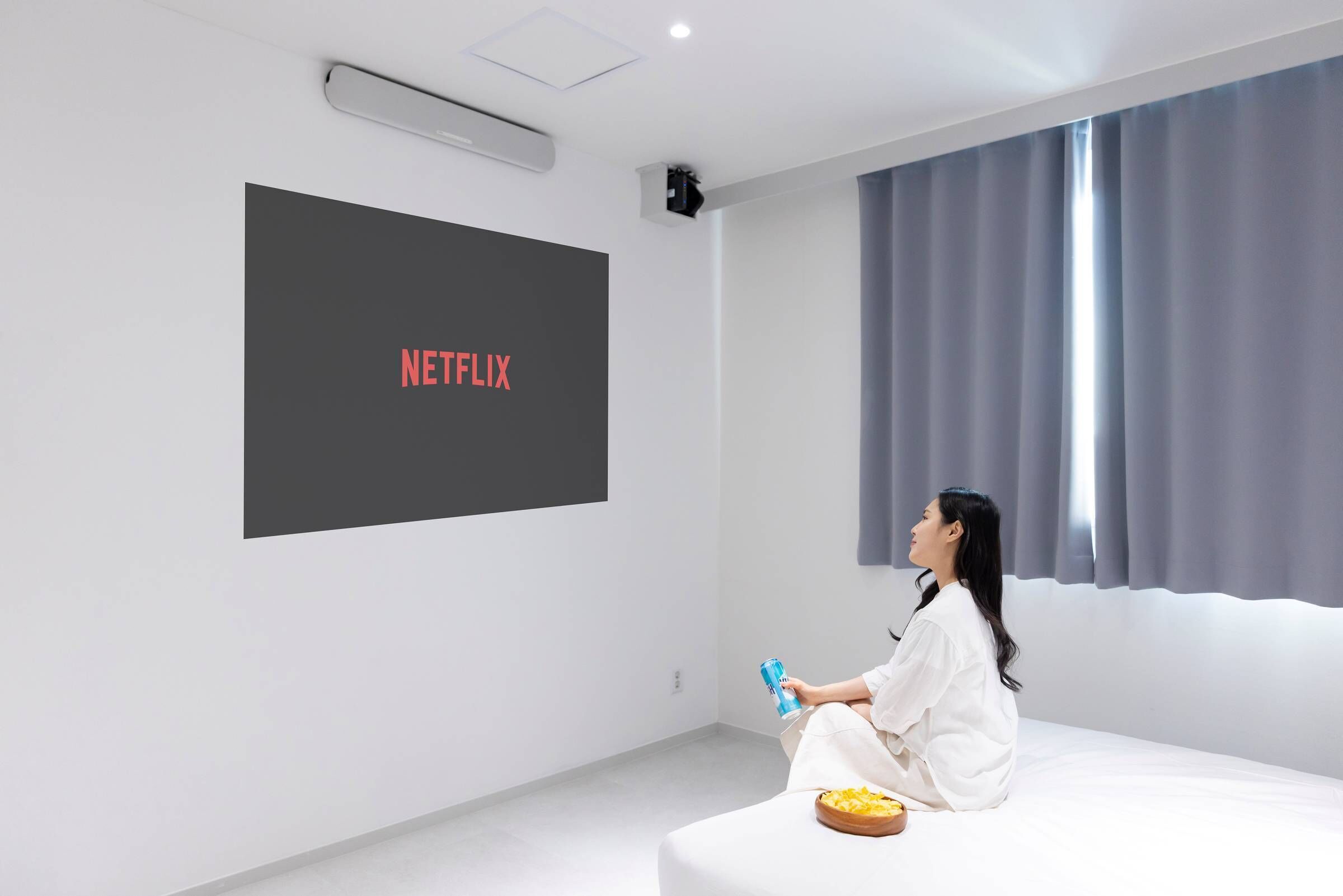 A modern hotel room with a large screen displaying the Netflix logo, a comfortable seating area, and a view of the outdoors through large windows.