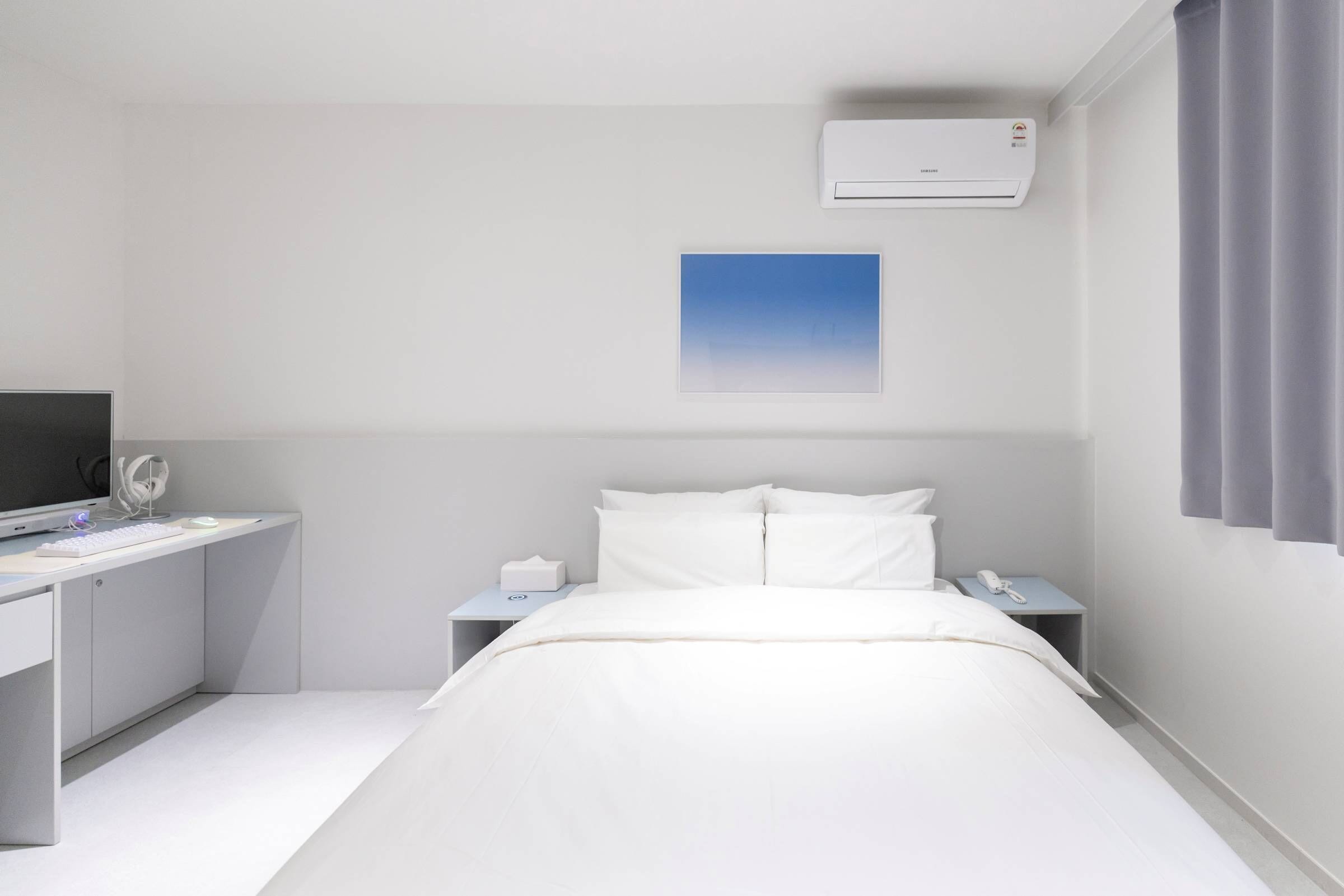A hotel room with a bed, a desk, a computer, a phone, and an air conditioner.