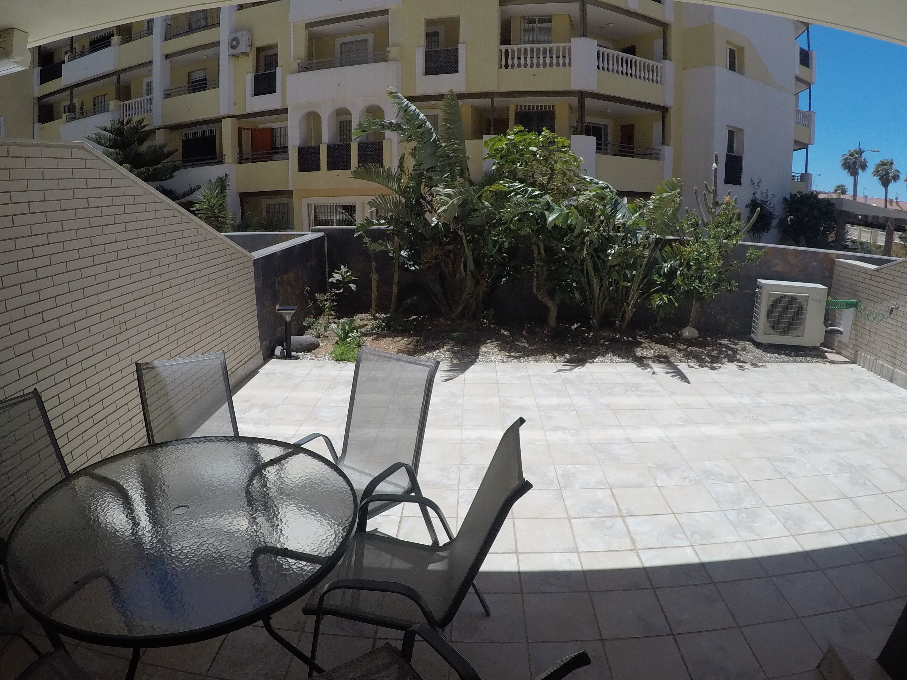 Une cour avec une table ronde en verre et des chaises, entourée d’un mur de briques et d’un bâtiment doté de balcons.