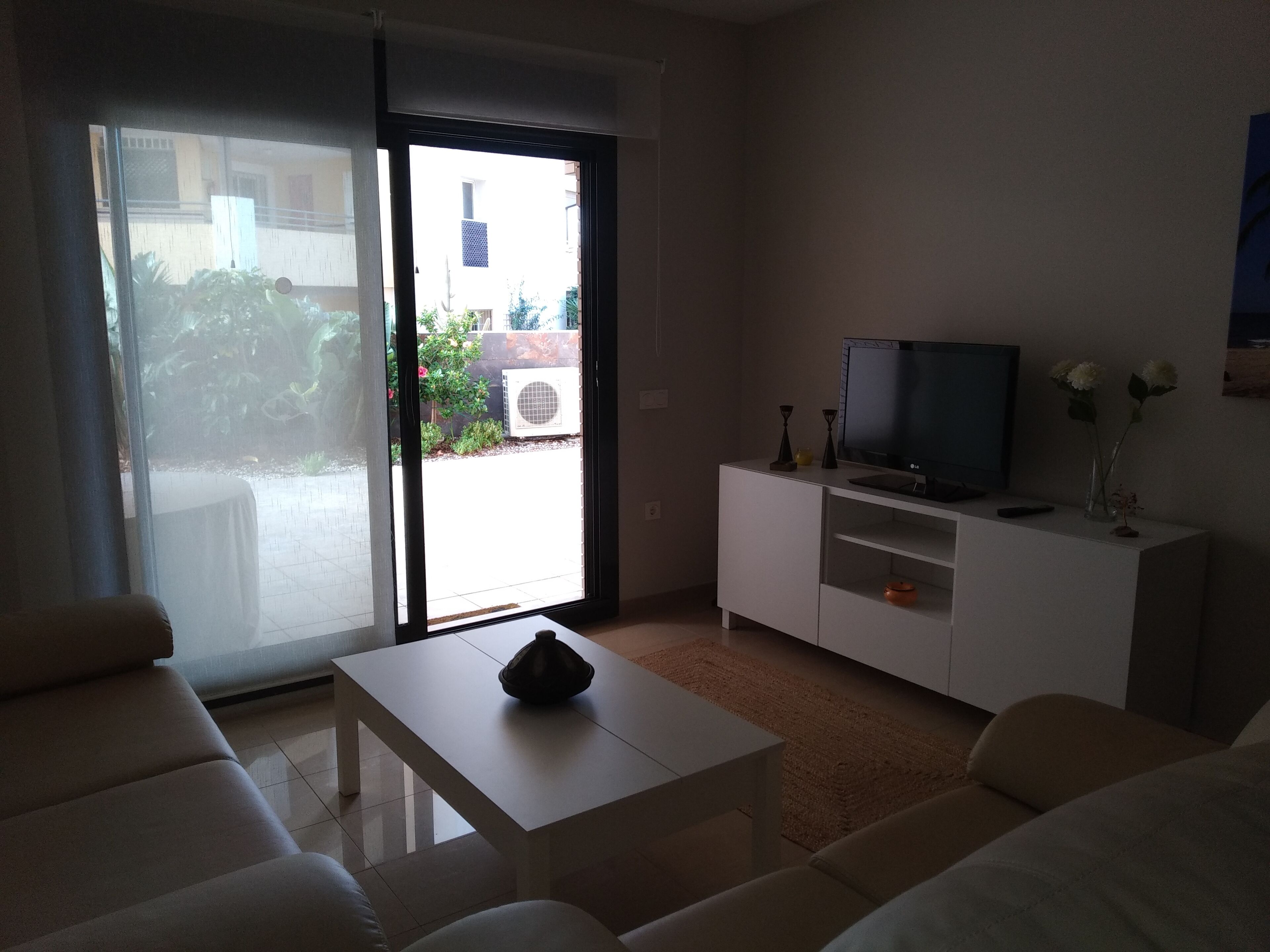 A living room with a sofa, a coffee table, a TV on a white cabinet, and a sliding glass door leading to a garden.