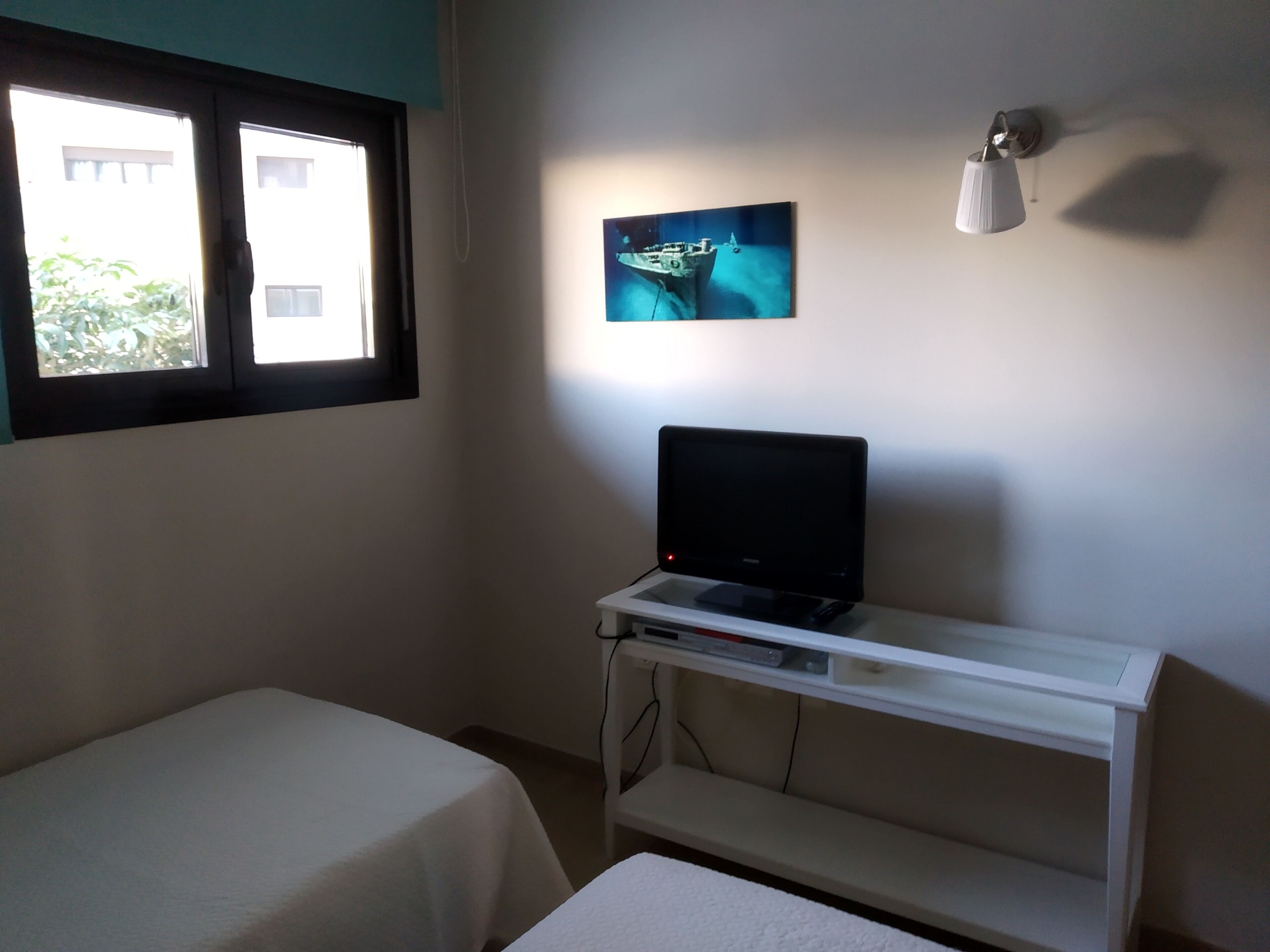 Ein Zimmer mit einem Bett, einem Schreibtisch mit Laptop, einer Wandlampe und einem Fenster mit Blick auf Gebäude und Grünflächen.