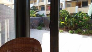 Une vue depuis un balcon, avec un fauteuil en osier, un sol carrelé et une cour intérieure agrémentée de plantes et d’un mur.