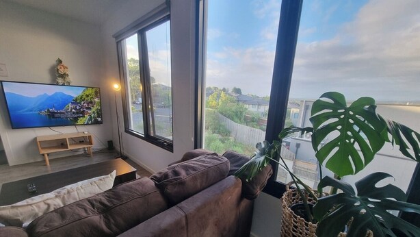 Sala de estar com sofá de couro marrom, televisão fixada na parede e uma janela grande com vista para uma área residencial.