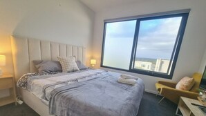 Quarto de hotel moderno com uma cama grande, duas luminárias de cabeceira, vista da cidade por uma janela ampla e uma poltrona pequena.