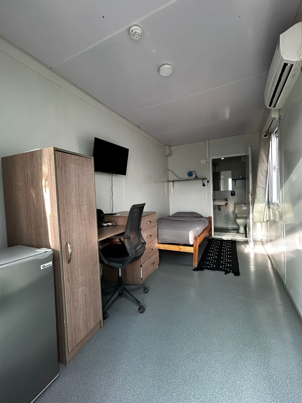 A compact room with a bed, a desk, a chair, a television, and an air conditioning unit.