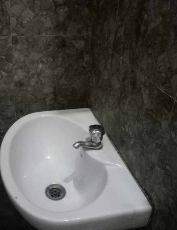 A bathroom with a white sink, a chrome faucet, and a drain. The walls are tiled with a dark, speckled pattern. - Serenity Space  (Yercaud)