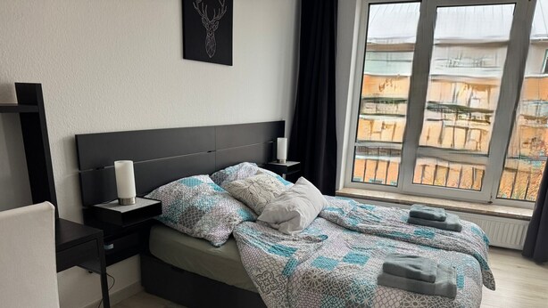 Un dormitorio moderno con una cama grande, una mesita de noche con lámpara, y vistas a un paisaje urbano a través de una ventana.