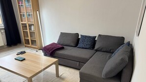Un salón moderno con un sofá gris, una mesa de centro de madera y vistas a un edificio a través de un ventanal.