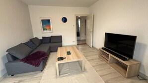 Una sala de estar con un sofá gris, una mesa de centro de madera, un televisor de pantalla plana sobre un soporte de madera y un cuadro en la pared.