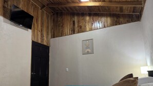 A room with a wooden ceiling, a bed with a blue bedspread, two pillows, a black door, and a wall-mounted TV. - Cabina Mandarina  (Provincia de Puntarenas)