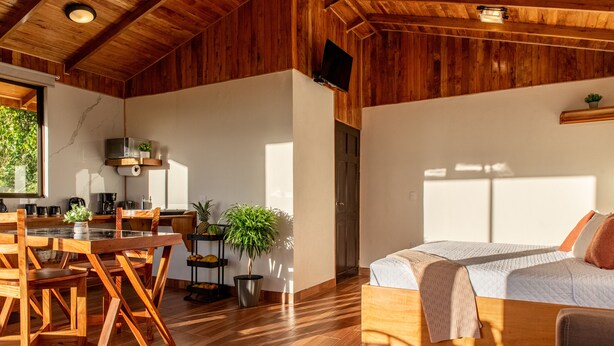 A spacious room with a wooden floor, a bed with a white quilt, a wooden dining table with chairs, and a kitchen area with a microwave and a plant. - Cabina Mandarina  (Provincia de Puntarenas)