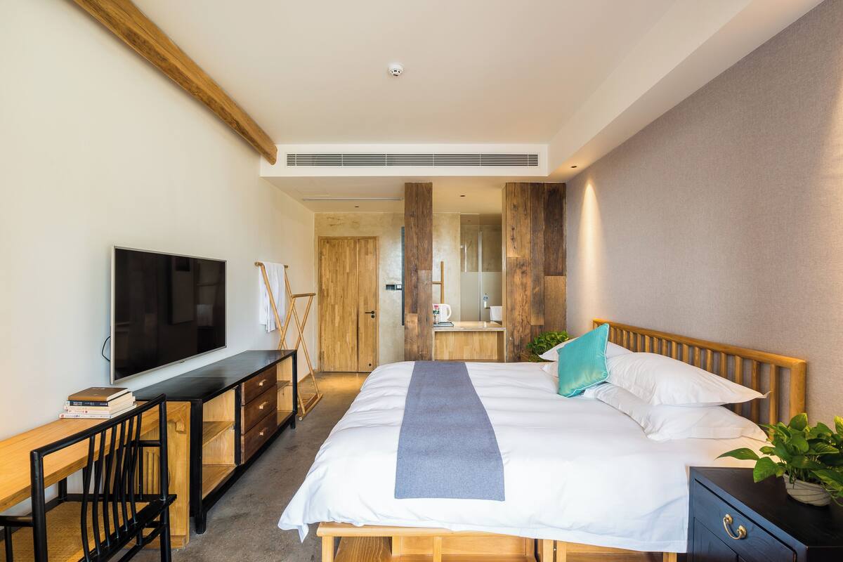 A modern hotel room with a large bed, a desk with a flat-screen TV, a wooden bench, and a small plant.
