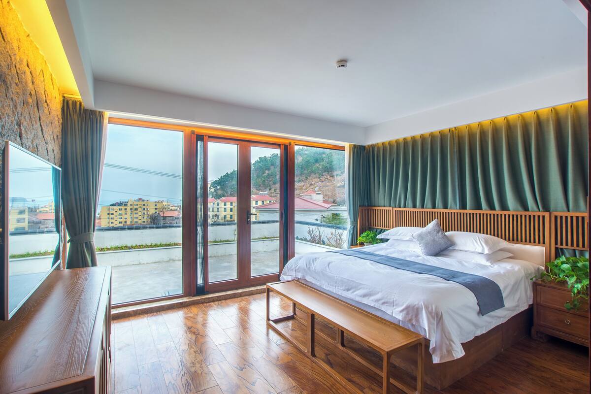 A modern bedroom with a large bed, wooden flooring, a bench, a nightstand, and a view of a cityscape through large windows.