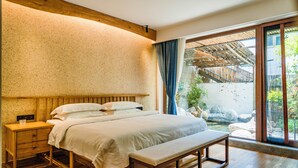 Un dormitorio con cabecera de madera, mesita de noche de madera, un banco y vistas a un jardín a través de una puerta corrediza de vidrio.