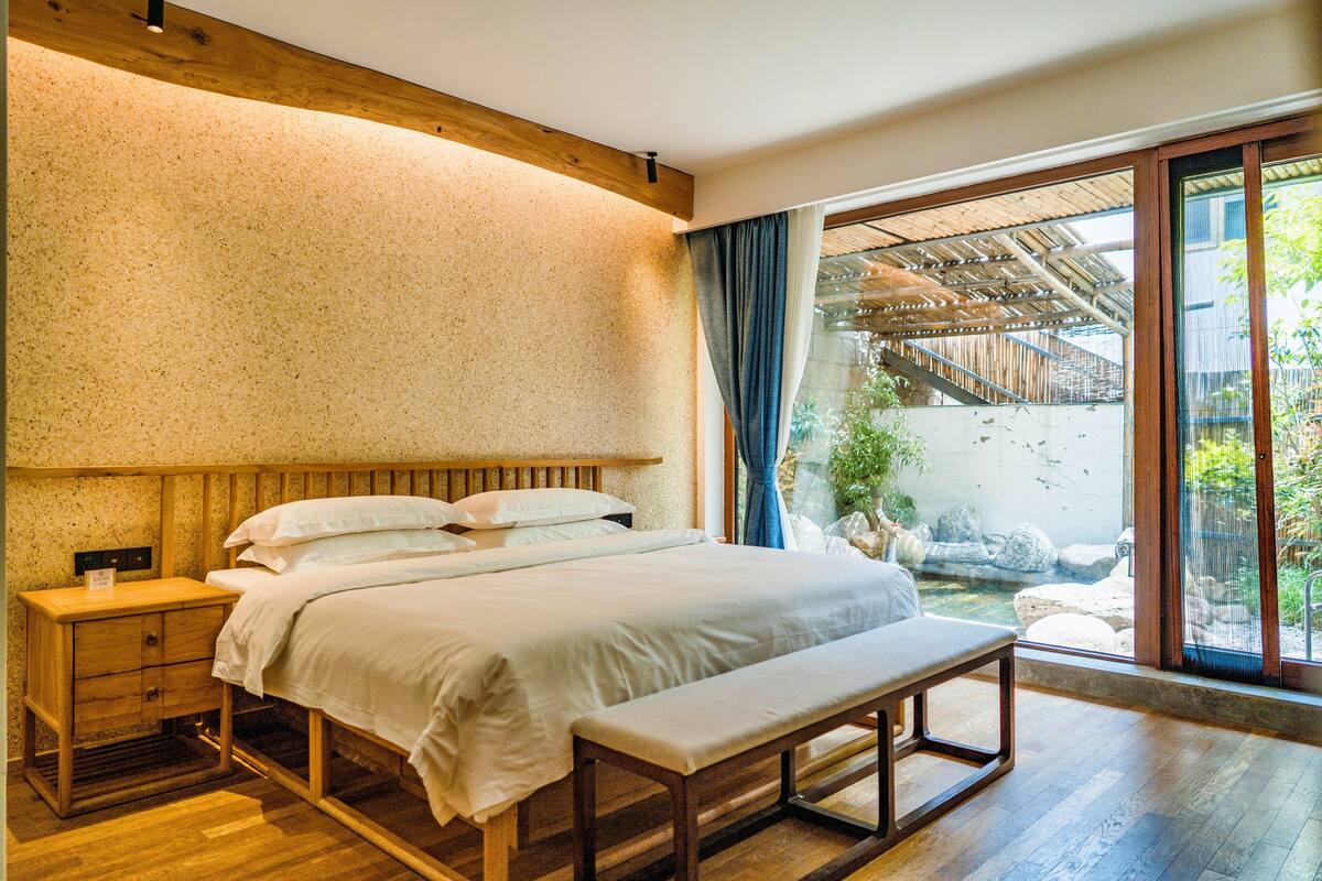 A bedroom with a wooden headboard, a wooden nightstand, a bench, and a view of a garden through a sliding glass door.
